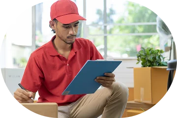 Bir teslimat görevlisi, taşınma kutuları arasında oturuyor ve envanter listesini kontrol ediyor; profesyonel taşımacılık ve lojistik hizmetlerini temsil ediyor.